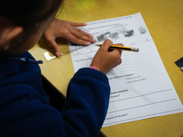 Child in a blue sweater writes on a French worksheet at a yellow desk, holding a pencil near the page.