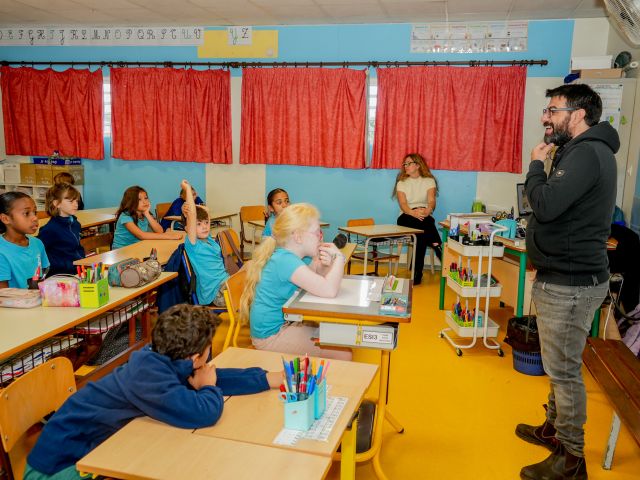 Bearded man in a dark hoodie speaks to a classroom full of young students seated at desks with blue walls and red curtains behind them.