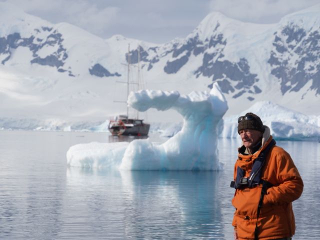 Man in an orange parka stands at a chilly shore with a blue iceberg arch and a sailing ship in the background.