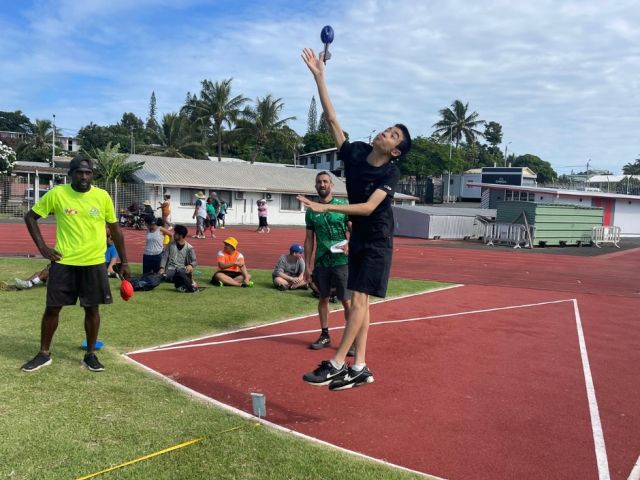 Two athletes leap to strike a blue ball during a track-side sport drill while teammates watch on a sunny day.