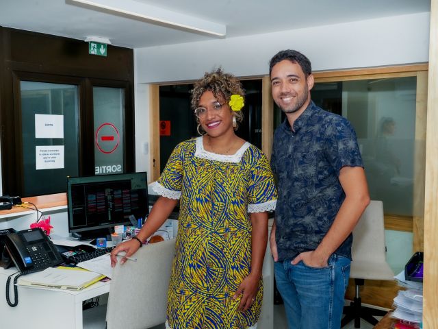 Two coworkers pose for a photo in an office reception area, the woman in a bright yellow patterned dress with a yellow flower in her hair and the man in a blue patterned shirt smiling beside her.