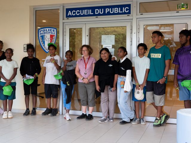 Group of teens and a woman posing outside a police station entrance, holding green caps. The door and a 'Accueil du Public' sign are visible behind them.
