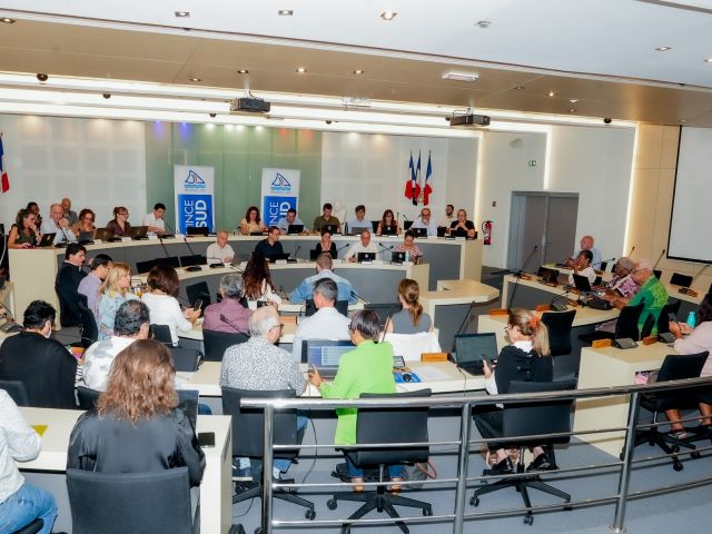 Large conference room with a panel of speakers at the front, French flags behind them, and attendees using laptops and notebooks.