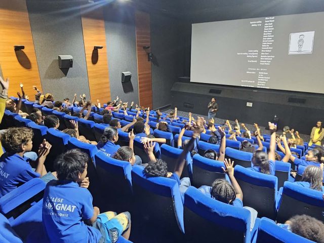 Large lecture hall filled with students in blue uniforms raising hands as a speaker presents at the front.