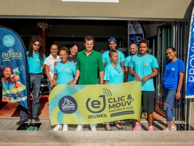 Group of diverse volunteers in turquoise shirts pose together behind a bright yellow banner at an outdoor event doorway, with Province Sud banners nearby.