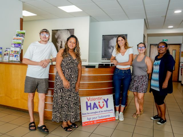 Group of five adults posing in a bright lobby near a curved wooden reception desk, with a 'Valentin Hauy' sign in front (charity event).