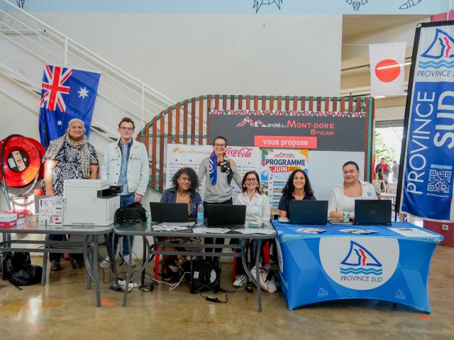 Group of six people standing and sitting behind a desk at an information booth, with banners reading Province Sud and Mont-Dore and various flags visible in the background (Australia flag on the left).